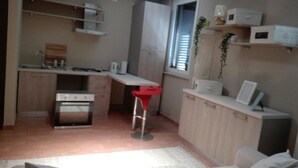 A compact kitchen with a stove, sink, and a red bar stool. There are cabinets and a window with blinds.