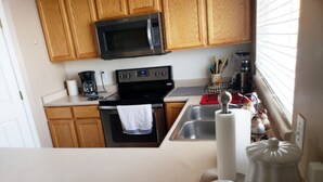 Une cuisine avec des meubles de rangement en bois, un four à micro-ondes, une cuisinière, un évier et une fenêtre avec des stores.