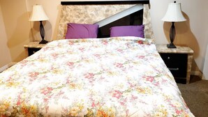 Un lit bien fait, recouvert d’une couvre-lit à motifs floraux, deux tables de chevet avec des lampes et une tête de lit dont la texture rappelle celle de la pierre.