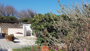 Un jardín con diversas plantas y flores, una terraza de madera y un edificio blanco con tejado de tejas.