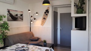 En moderne stue med en sofa, et sofabord med et mønstret dug, en gulvlampe, en surfbræt-dekoration og et fjernsyn på et stativ.