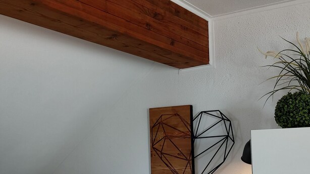 Ein modernes Wohnzimmer mit sichtbarem Holzbalken, einem geometrischen Wandkunstwerk, einer weißen Lampe, einem dunklen Sofa mit gemusterten Kissen und einem kleinen Topfpflanzen.
