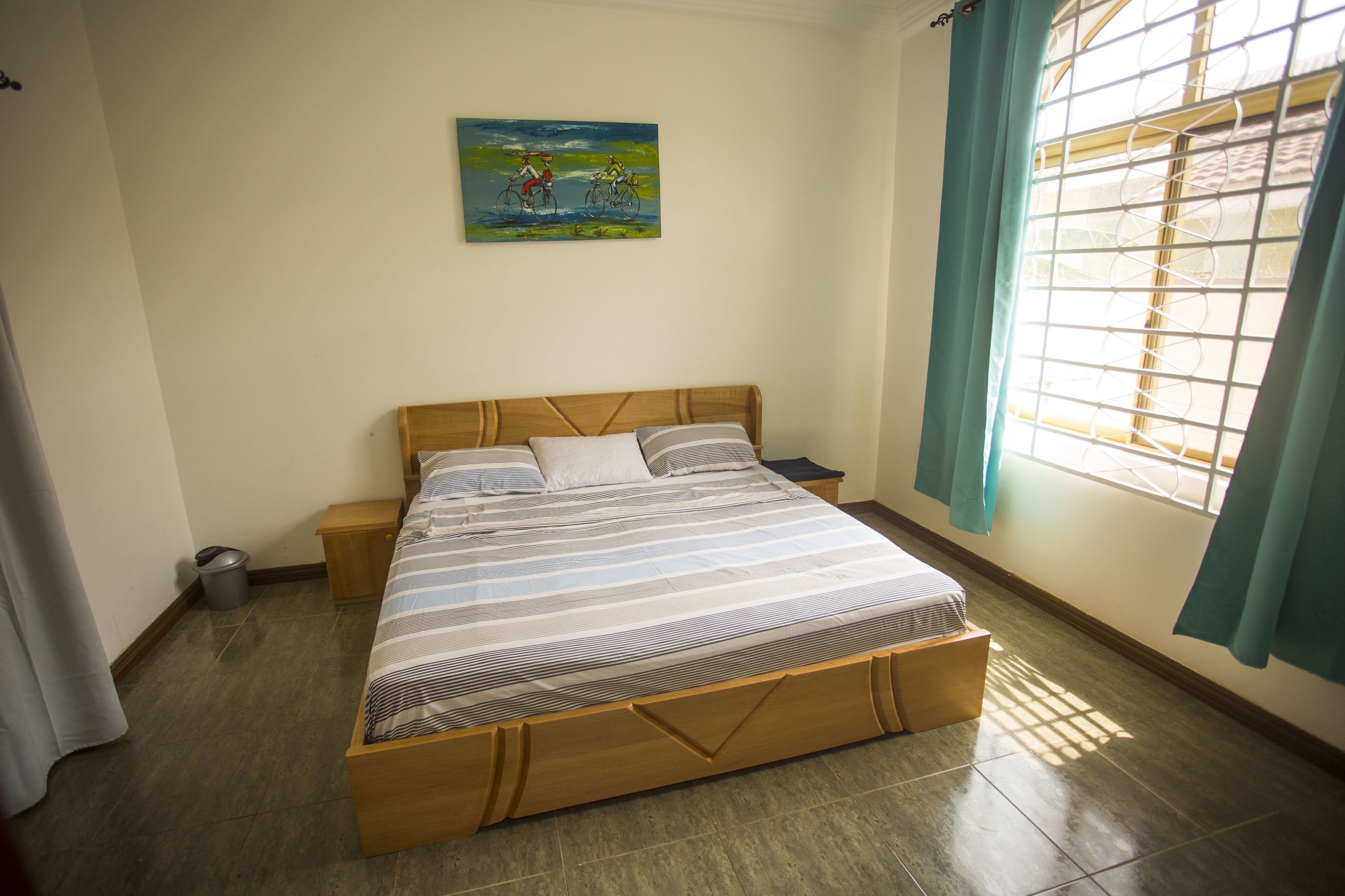 A bedroom with a wooden bed, a nightstand, a trash bin, a window with curtains, and a painting on the wall.
