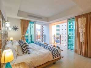 Relax in a spacious room with a large bed, bedside lamp, and balcony seating area offering a view of the surrounding buildings.
