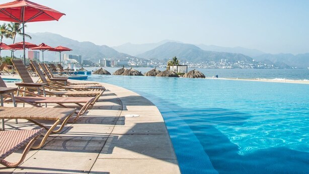 A poolside area with lounge chairs and umbrellas, a clear view of the ocean and mountains, and a cityscape in the background. - BEACHFRONT & TERRACE CONDO! (Puerto Vallarta)