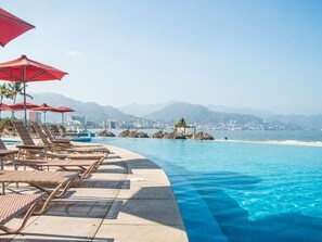 Een zwembadterras met ligbedden en parasols, met een helder uitzicht op de oceaan en bergen, en een stadszicht op de achtergrond.