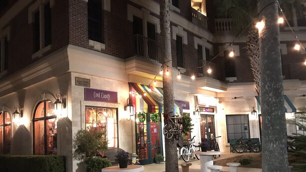 Escena nocturna de una calle con un edificio que muestra un letrero 'Cool Candy', mesas al aire libre y bicicletas.