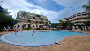 Zona de piscina con sillones de descanso y sombrillas, rodeada de edificios con balcones y palmeras.