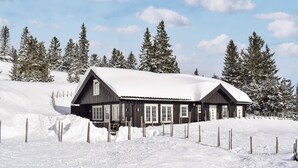 Eine Holzhütte mit schneebedecktem Dach, umgeben von schneebedeckten Bäumen und einem Zaun.