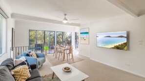 A spacious living area with a balcony, a dining set, a sofa, and a TV displaying a beach scene.