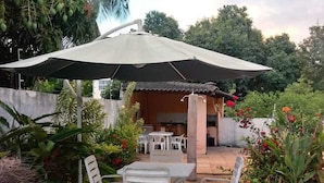 An outdoor patio with a table and chairs, a large umbrella, and a small building with a tiled roof. - House on Itaparica Island - Ponta de Areia Beach (Itaparica)