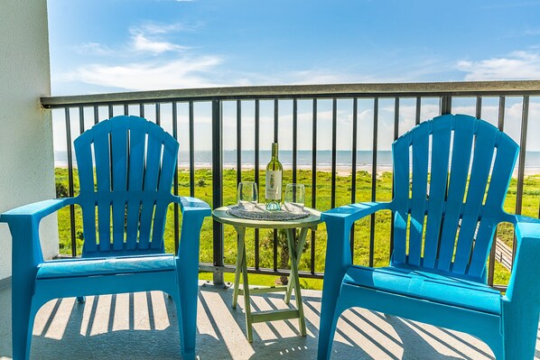 Deux chaises bleues et une petite table sur laquelle se trouvent une bouteille et des verres, le tout sur un balcon donnant sur la plage.