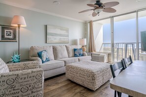 A cozy living room with a beige sofa, ottoman, and chairs, a dining table, and a large window offering a view of the ocean. - Sunny beachfront condo (Galveston)