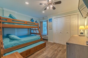 Un lit superposé avec une structure en bois et une literie bleue, un ventilateur de plafond, un bureau avec une lampe et une armoire.