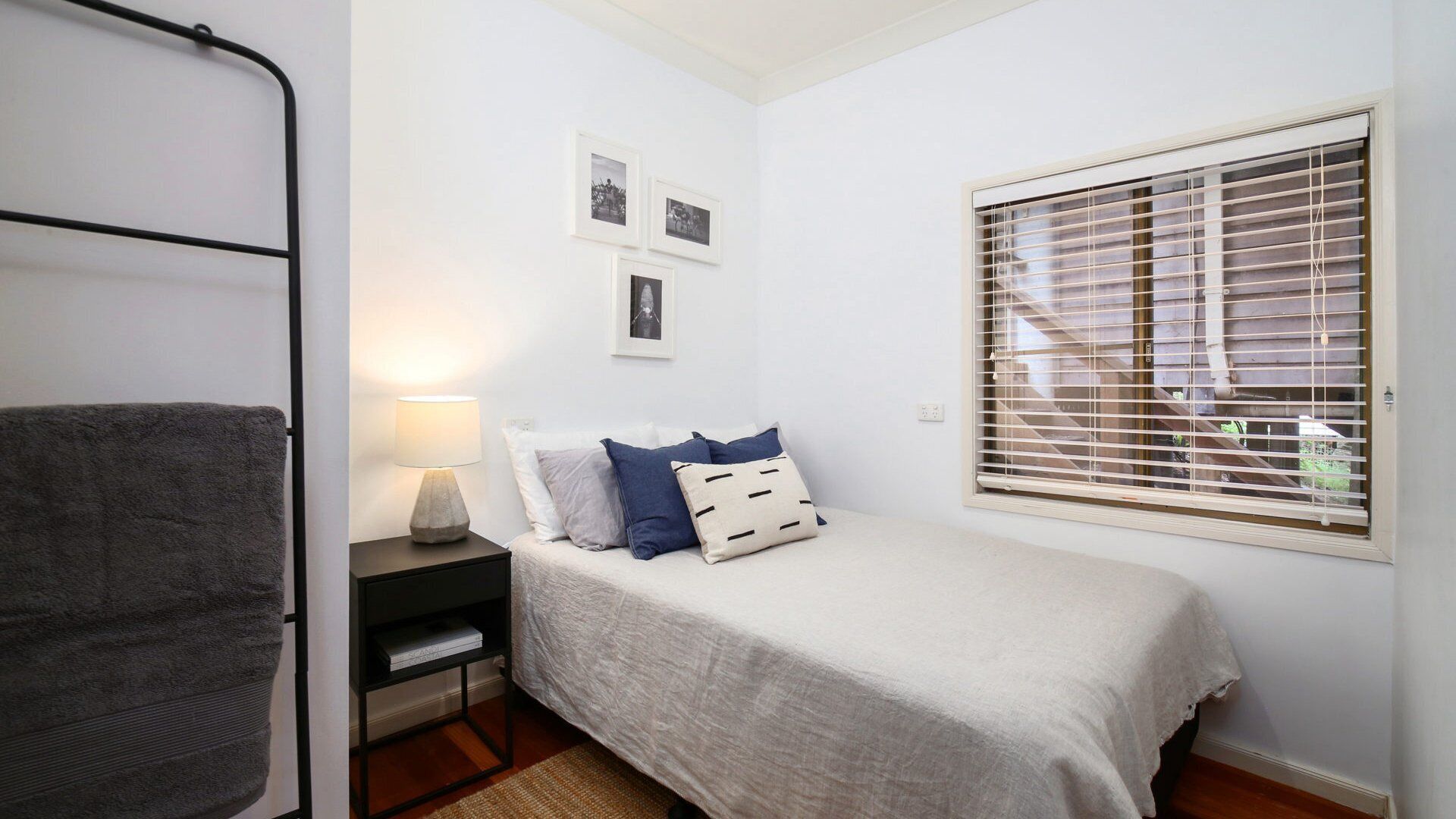 A bedroom with a bed, a nightstand, a lamp, a window with blinds, and a towel rack.
