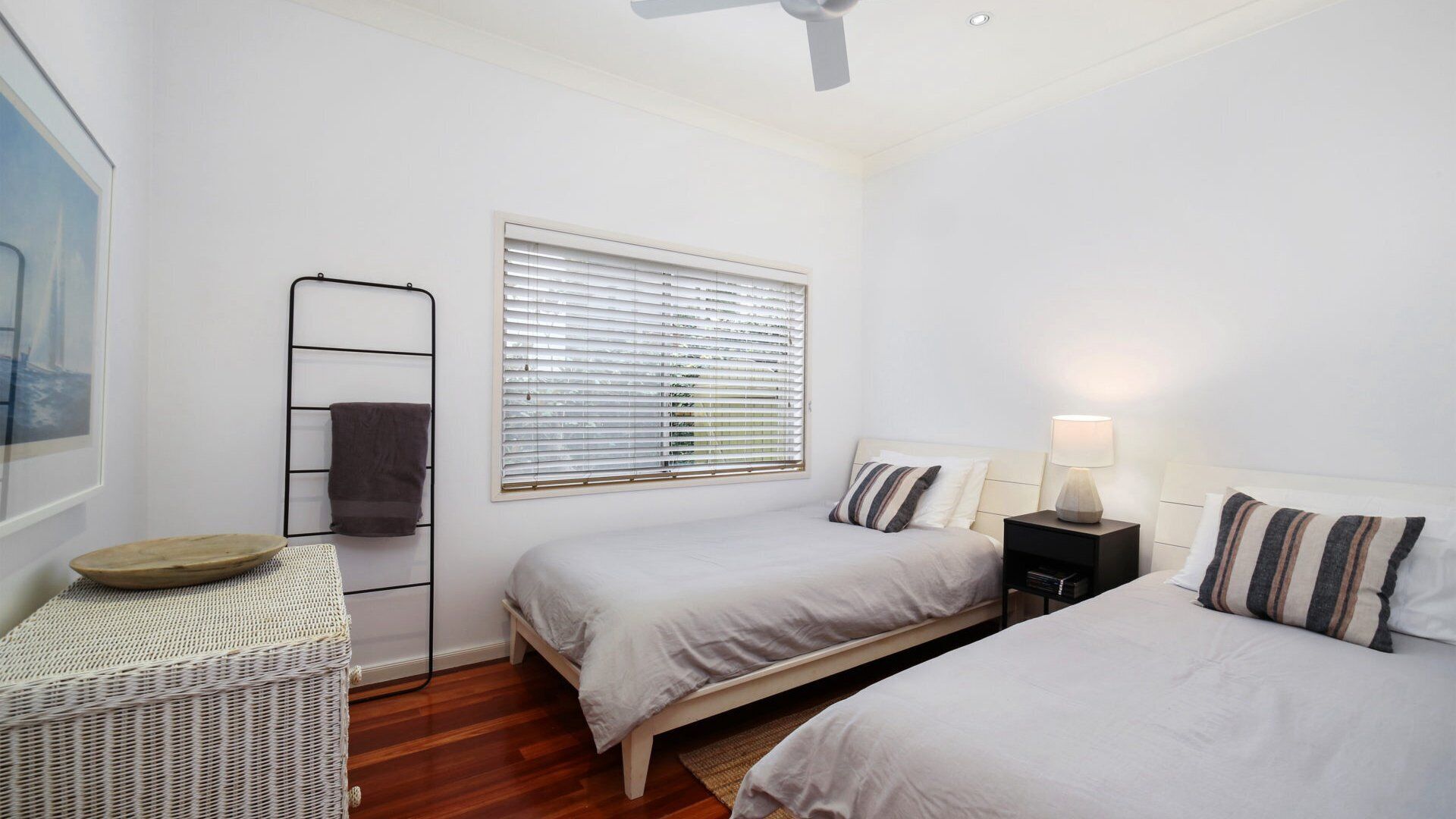 A bedroom with two beds, a ceiling fan, a window with blinds, a bedside table with a lamp, and a wicker chair.