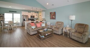 A spacious living room with a high ceiling fan, a dining area with a table and chairs, and a kitchen in the background. - You've earned the right to enjoy the best! (Bolivar Peninsula)