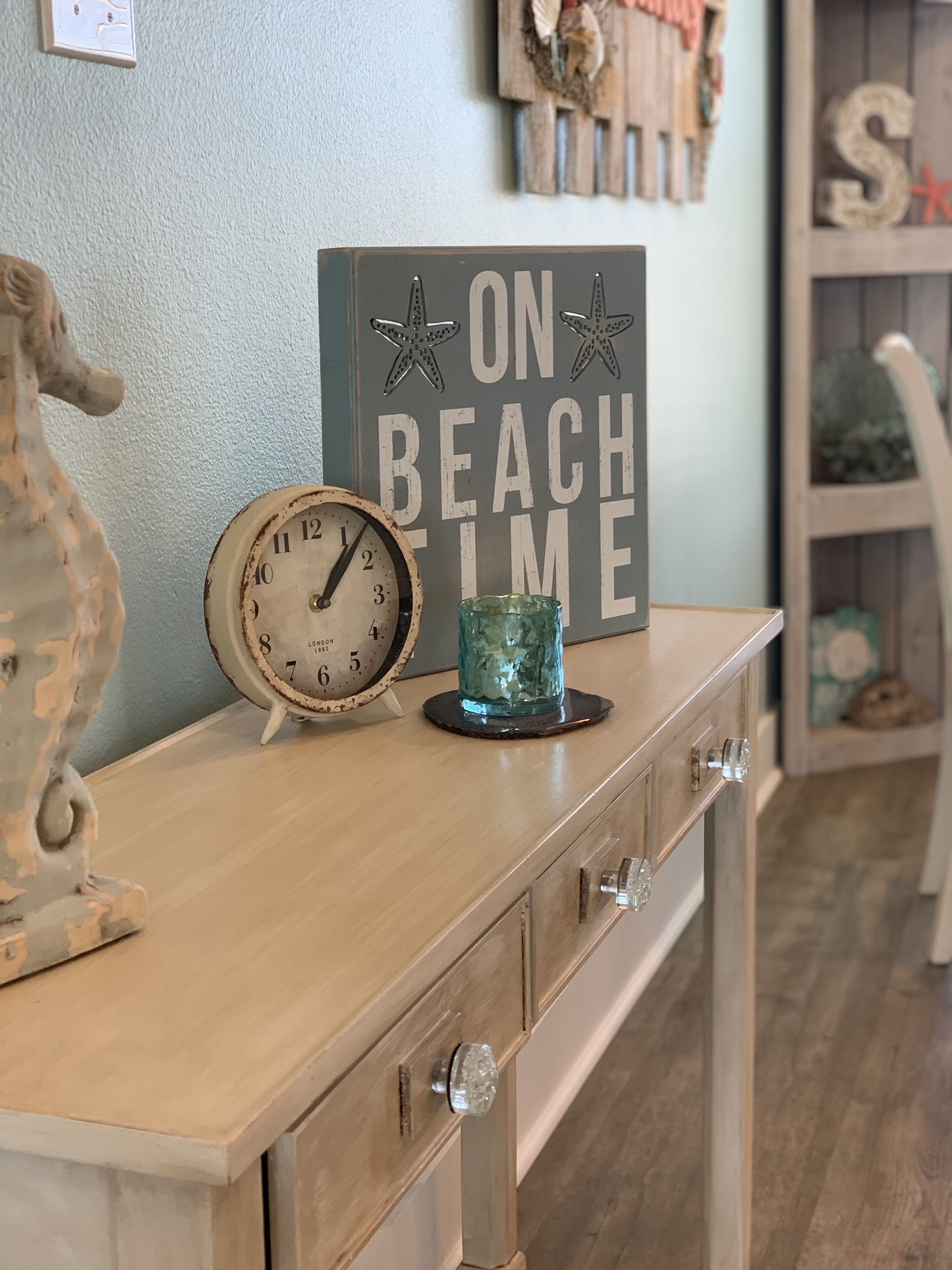 Une commode en bois, une horloge ancienne, un vase décoratif et une pancarte sur laquelle on peut lire : « EN MODE DÉTENTE À LA PLAGE ».