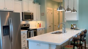 A modern kitchen with white cabinetry, stainless steel appliances, a large island with bar stools, and pendant lighting. - You've earned the right to enjoy the best! (Bolivar Peninsula)