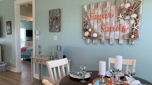 A dining area with a wooden table set for four, white chairs, and a wall decoration with the text "Sugar Sands". - You've earned the right to enjoy the best! (Bolivar Peninsula)