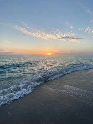 A beach with waves gently lapping onto the shore, a clear sky, and the sun setting on the horizon.