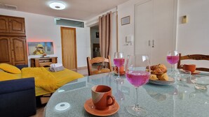 Un salon chaleureux avec un canapé, une table en verre sur laquelle sont disposées des boissons et des pâtisseries, et un téléviseur posé sur un meuble.