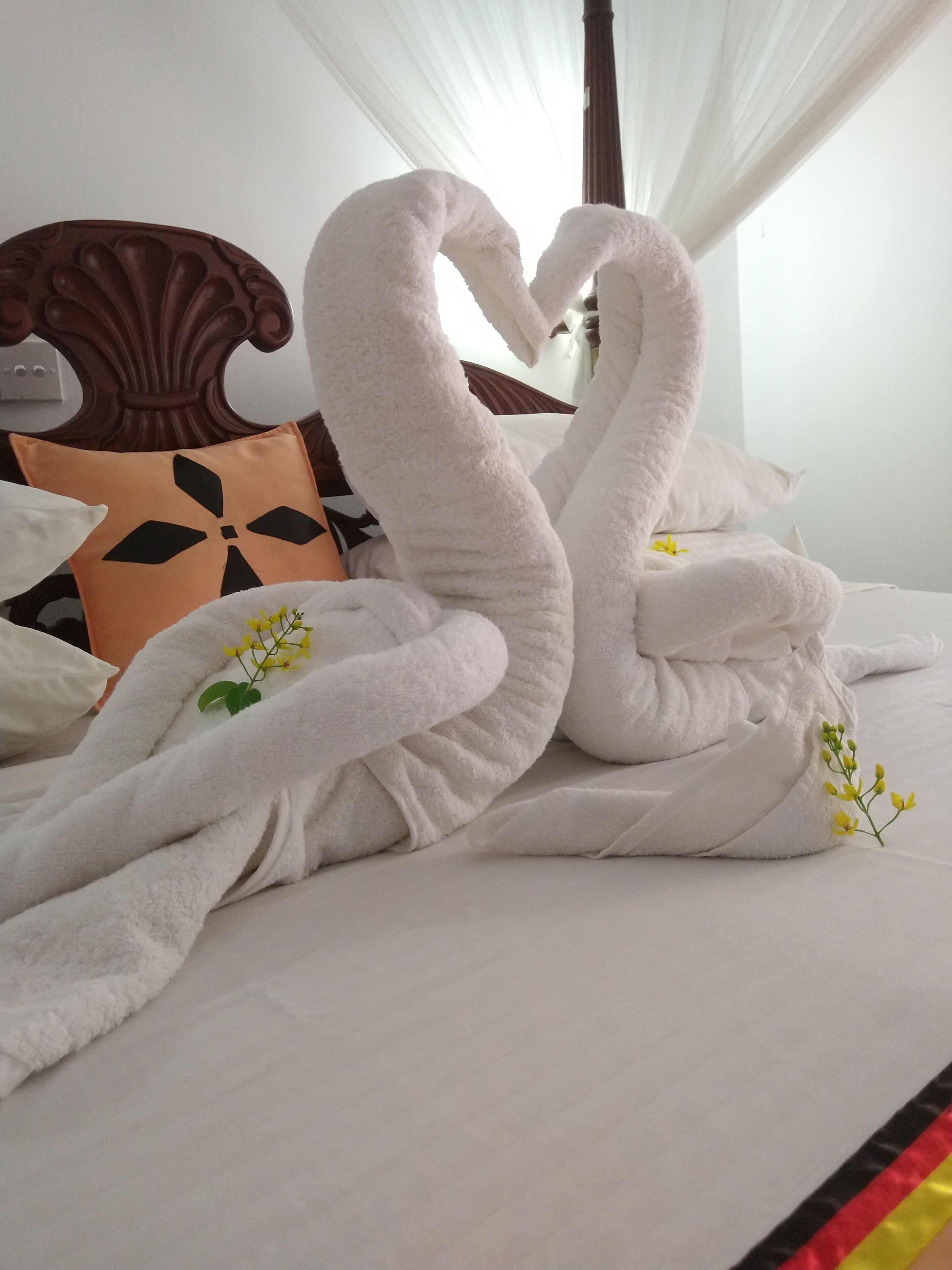 A bed with two large white towels arranged in the shape of swans, a brown headboard, and a yellow pillow with a black design.