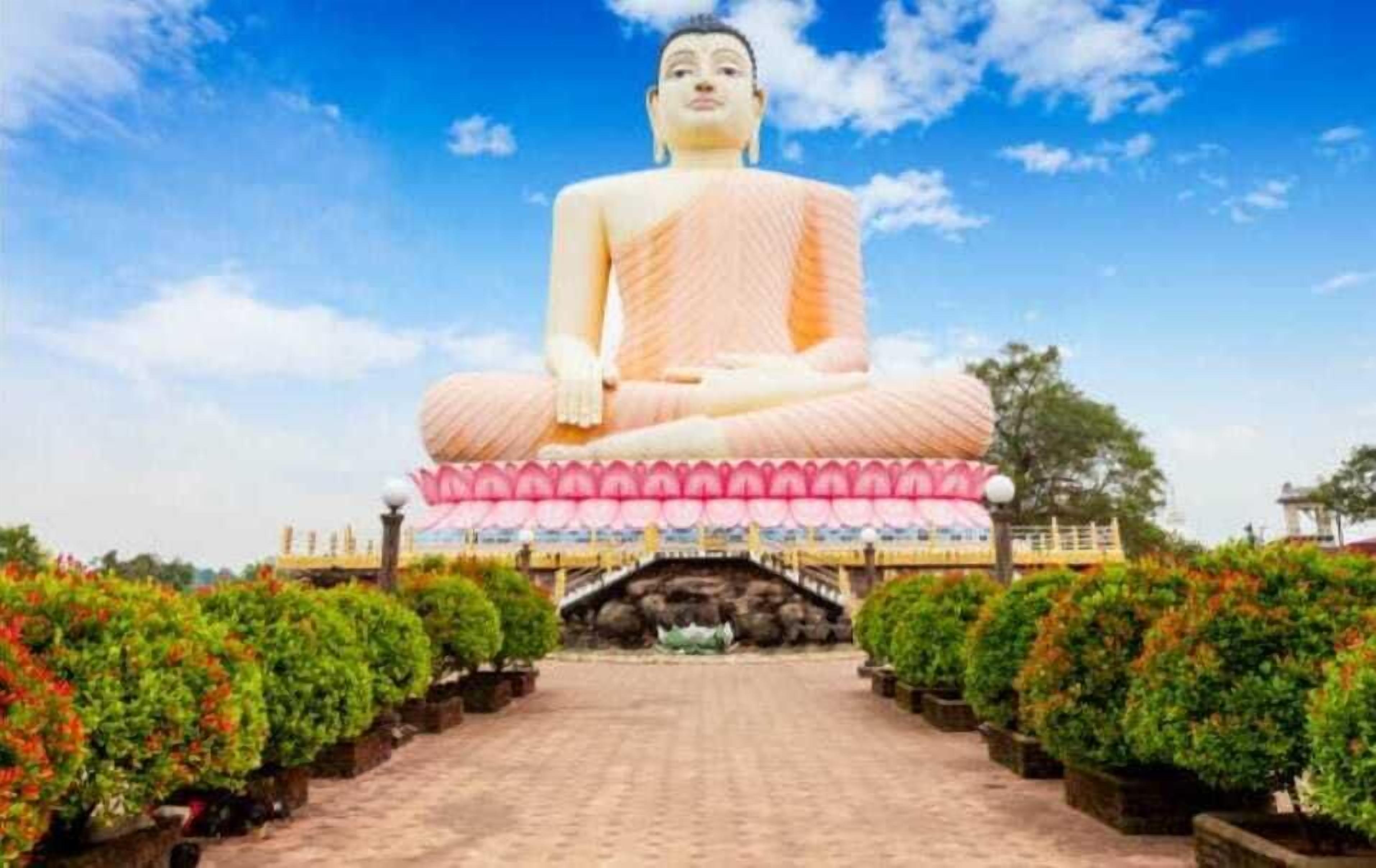 Una gran estatua de Buda sentado, sobre un pedestal de loto, rodeado de follaje y un cielo despejado.