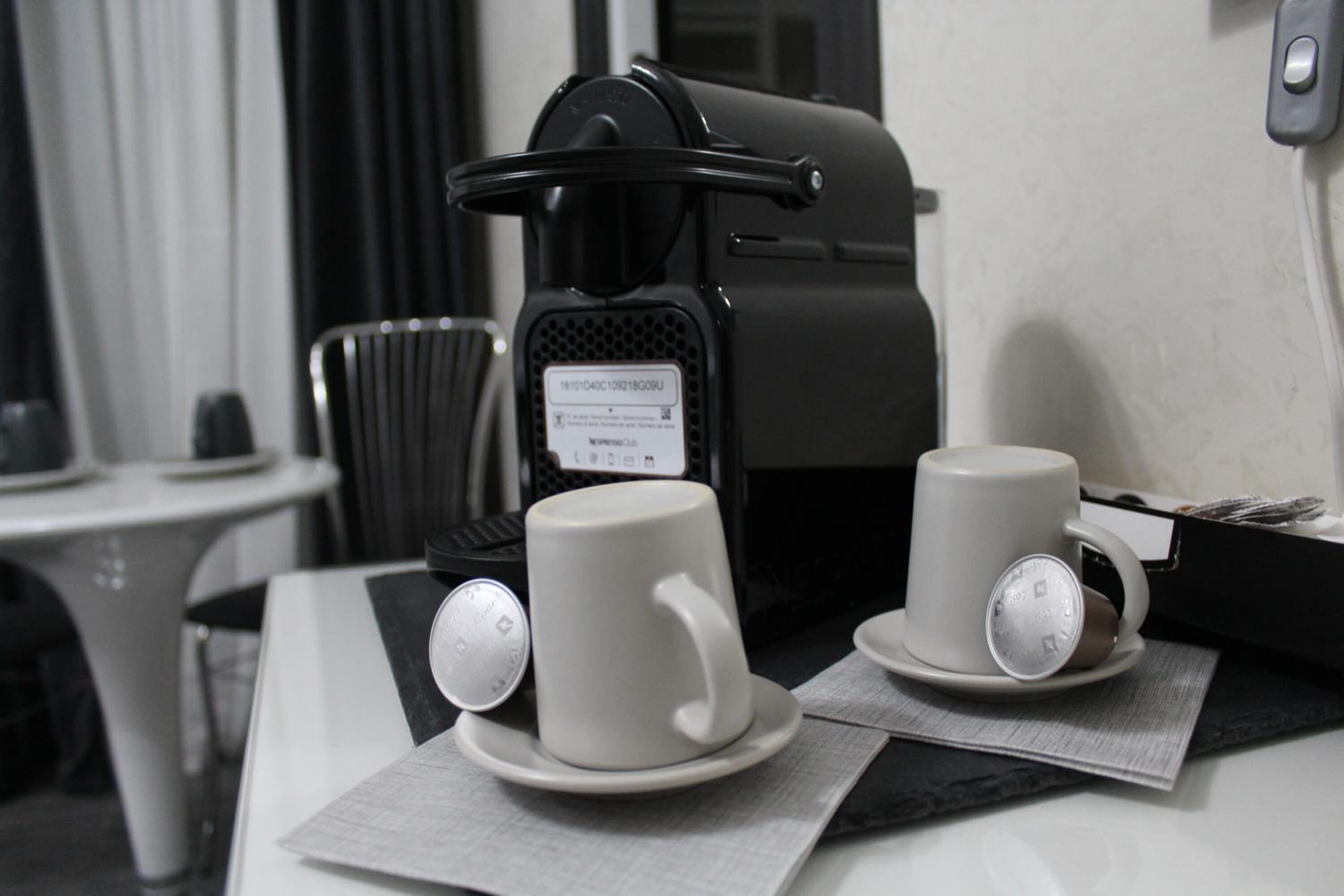 Una cafetera con dos tazas blancas sobre una mesa, una silla y una mesita con mantel blanco.