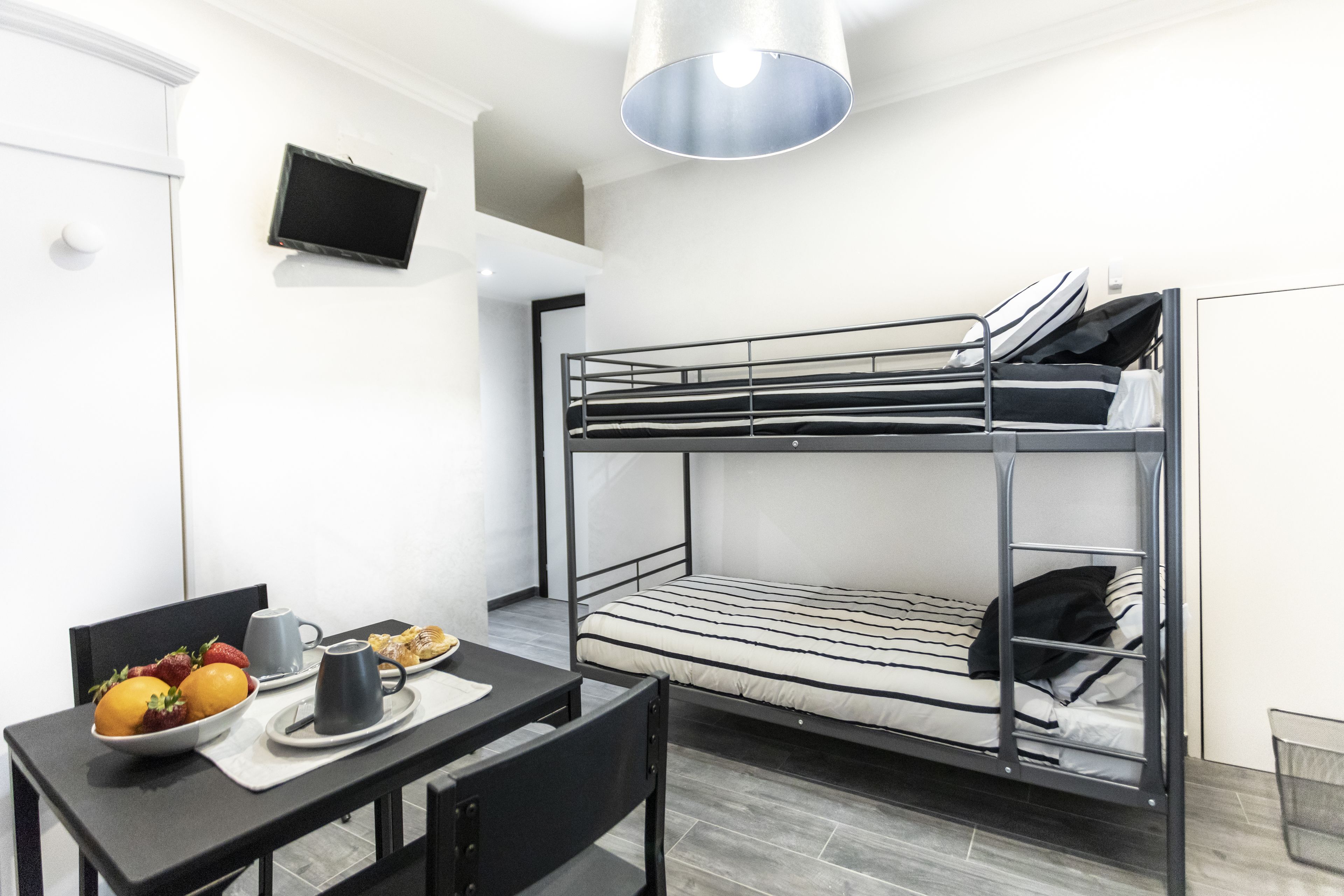 A bunk bed with a TV mounted on the wall, a table with a bowl of fruit and a mug, and a trash bin.