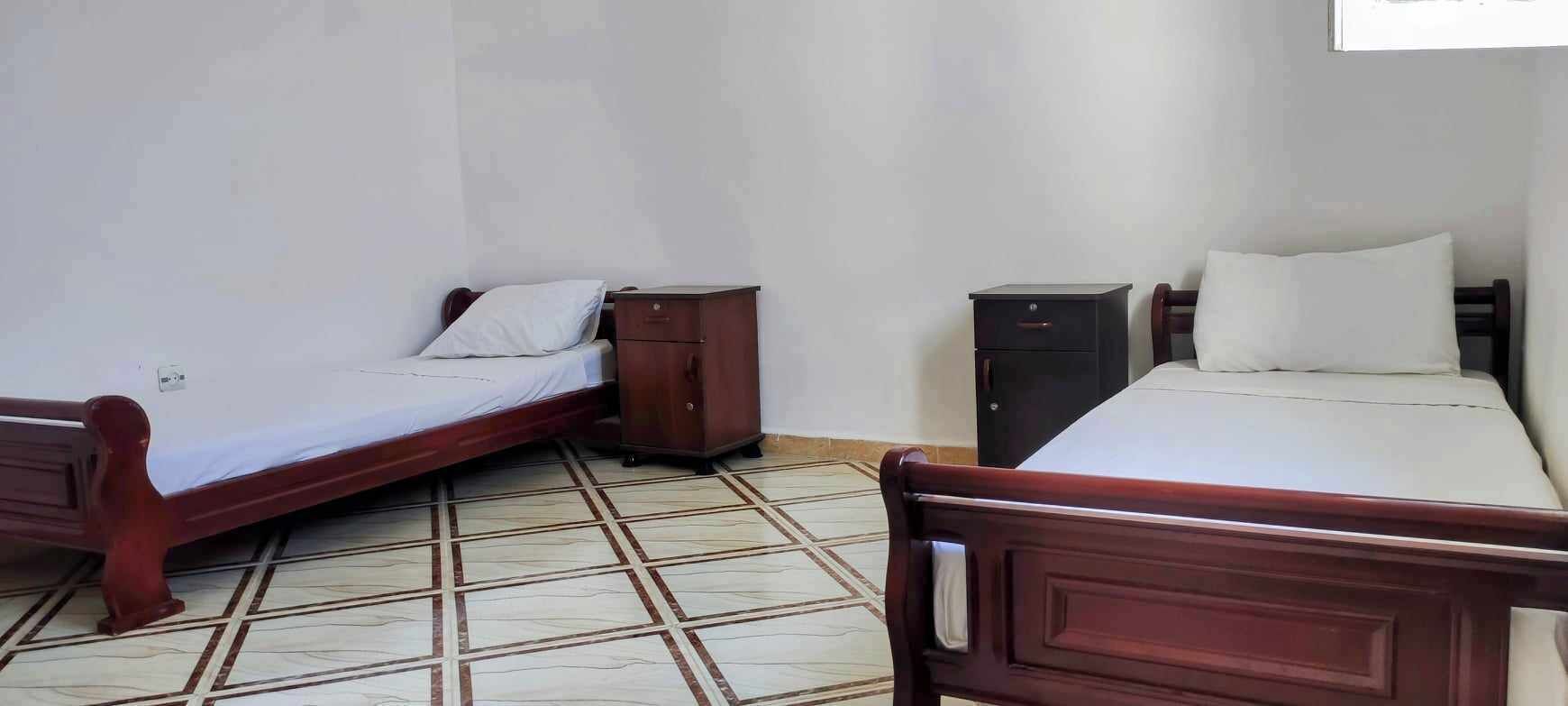 Une pièce avec deux lits en bois, chacun doté d’un matelas et d’un oreiller blancs, et une petite table de chevet en bois placée entre les deux. Le sol est recouvert de carreaux formant un motif.