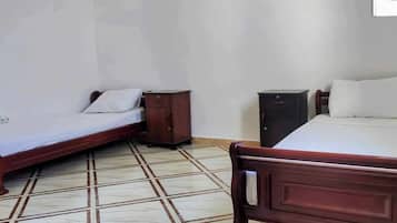 Une pièce avec deux lits en bois, chacun doté d’un matelas et d’un oreiller blancs, et une petite table de chevet en bois placée entre les deux. Le sol est recouvert de carreaux formant un motif.