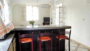 A kitchen with a granite countertop, three wooden chairs, a small island with a vase, and a television. - La Résidence Location Daoud (Souk El Tenine)