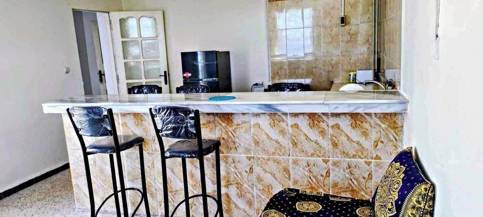 A kitchen with a marble countertop, two bar stools, a refrigerator, and a window with a view.