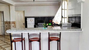 A kitchen with a marble countertop, three wooden bar stools, a refrigerator, a stove, and a window with a white curtain. - La Résidence Location Daoud (Souk El Tenine)