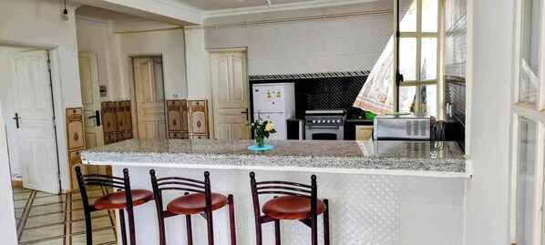 Una cocina con encimera de granito, refrigerador, estufa y lavamanos. Hay tres taburetes frente a la encimera.
