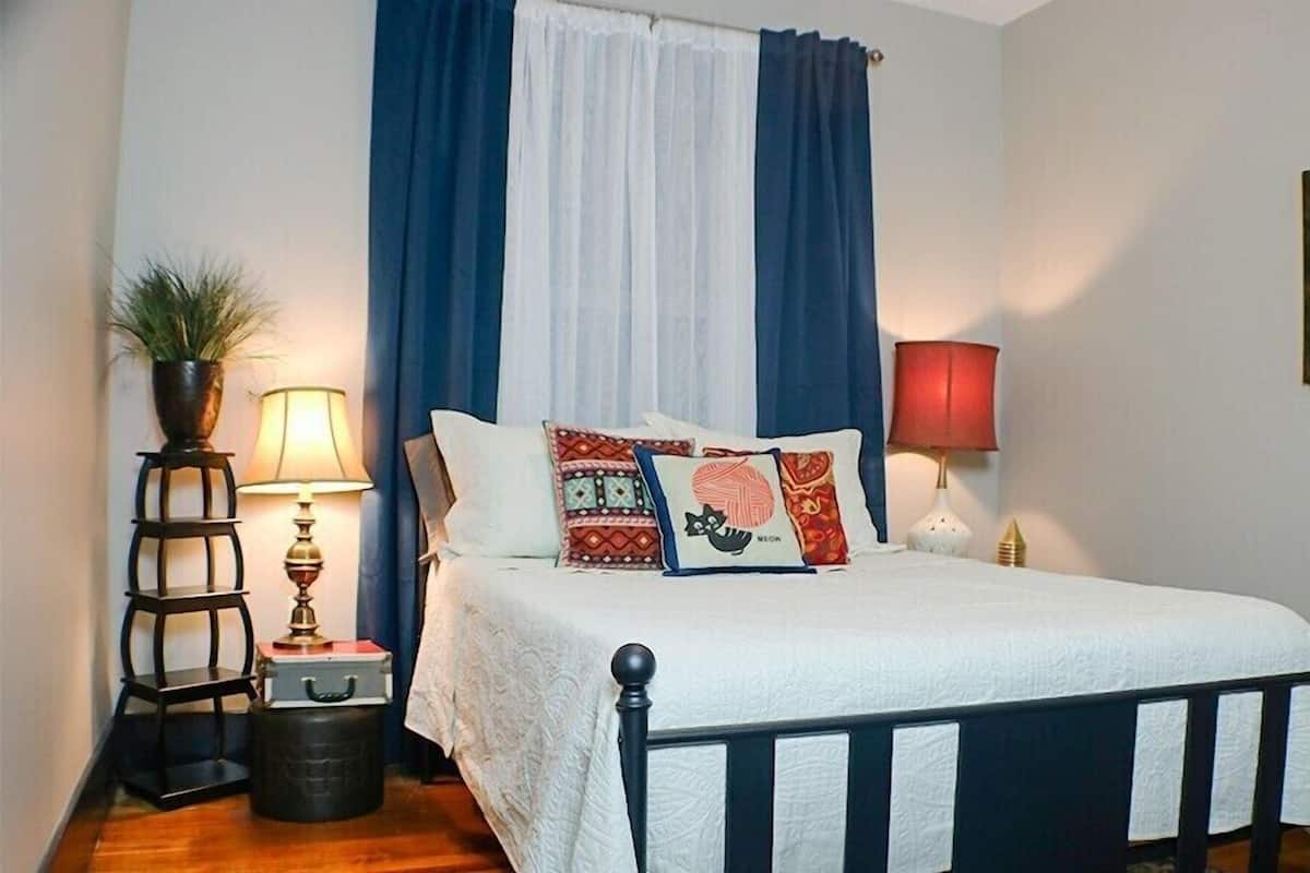 Una cama bien hecha con almohadones estampados, una lámpara de noche, una mesita con una planta y un cuadro en la pared.