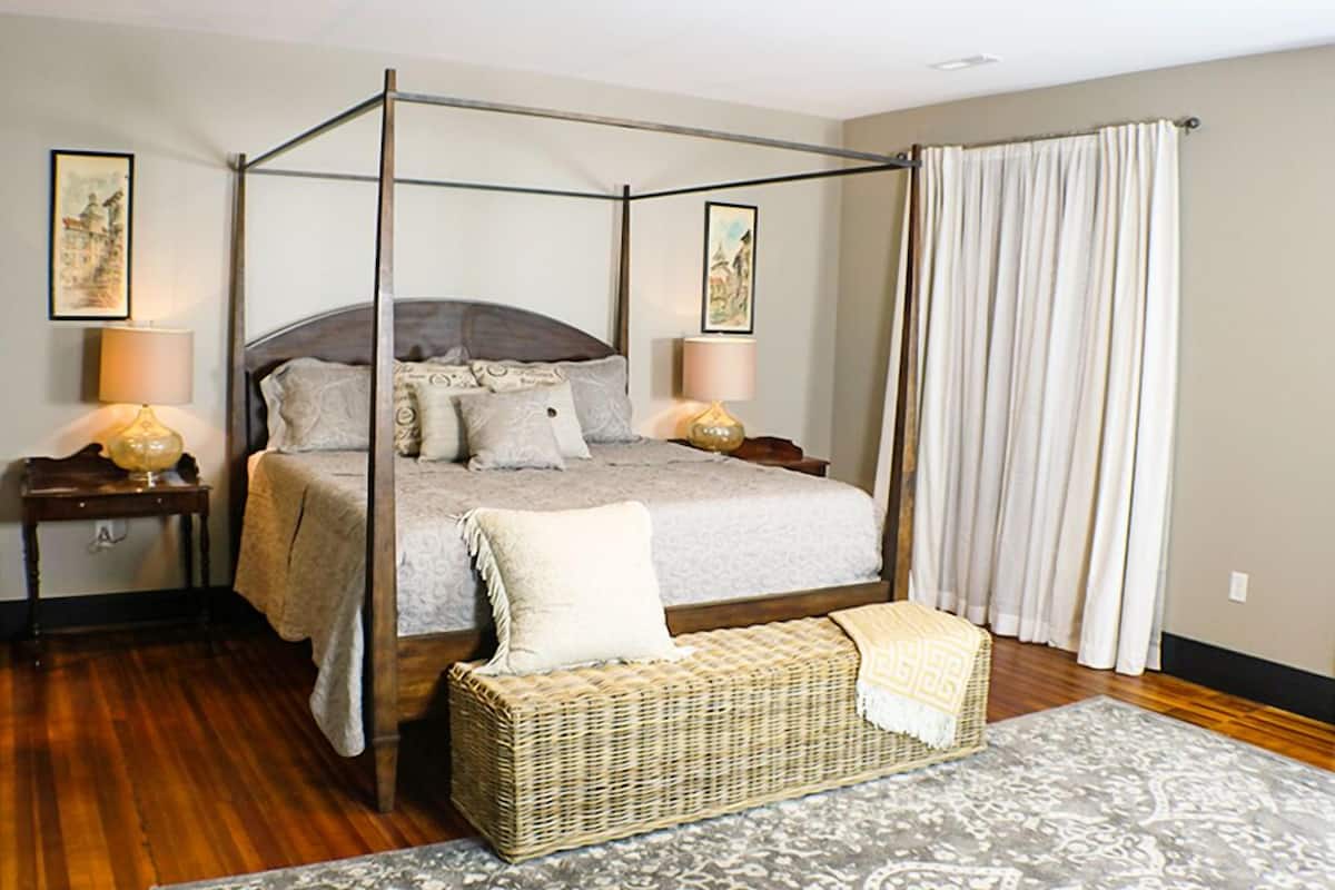 Una cama con dosel y cuatro columnas, con una colcha gris y varias almohadas, flanqueada por dos mesitas de noche con lámparas, un banco de mimbre y una alfombra estampada.