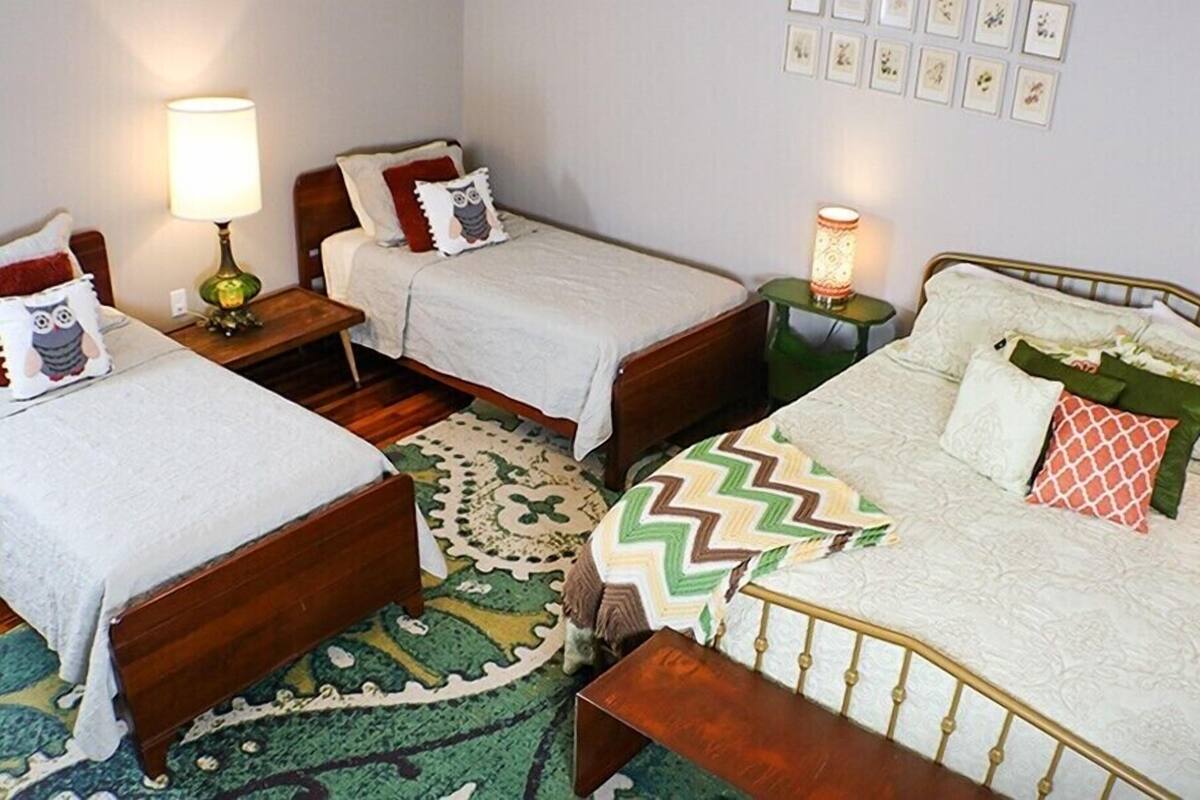 Una habitación con tres camas individuales, cada una con diferente estilo de ropa de cama y almohadas. Hay mesitas de noche con lámparas, una alfombra estampada en el suelo y una pared con cuadros enmarcados.