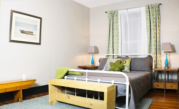 A bedroom with a bed, bedside tables, a nightstand, a window with curtains, a framed picture on the wall, and a wooden bench.