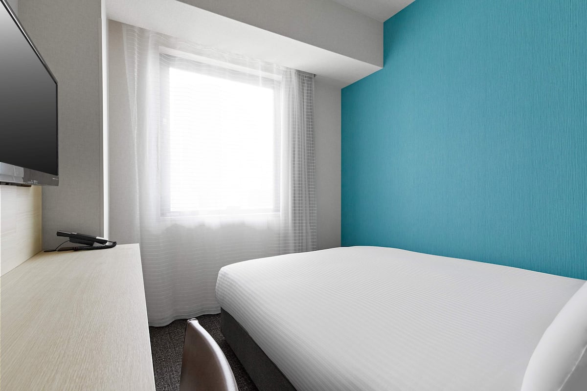 A hotel room with a bed, a desk, a television, and a window with sheer curtains.