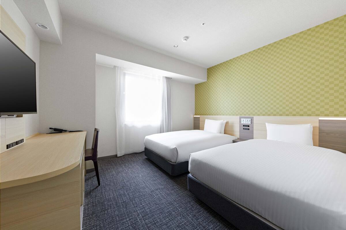 A hotel room with two beds, a desk, a TV, and a window with curtains.