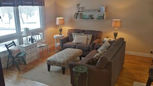 A cozy living room with a sofa, ottoman, and a side table with a lamp. There is a chair with a cushion, a small table with a lamp, and a shelf with decorative items. - Charming and comfortable home away from home (Rapid City)