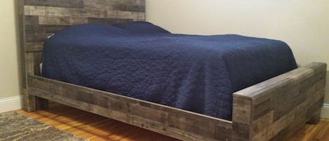 A wooden bed with a dark blue quilt in a room with a wooden floor and a gray rug.