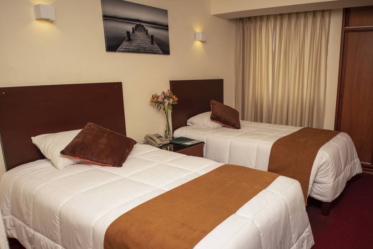 A hotel room with two beds, each with a brown throw blanket, white bedding, and a nightstand with a phone and vase of flowers.