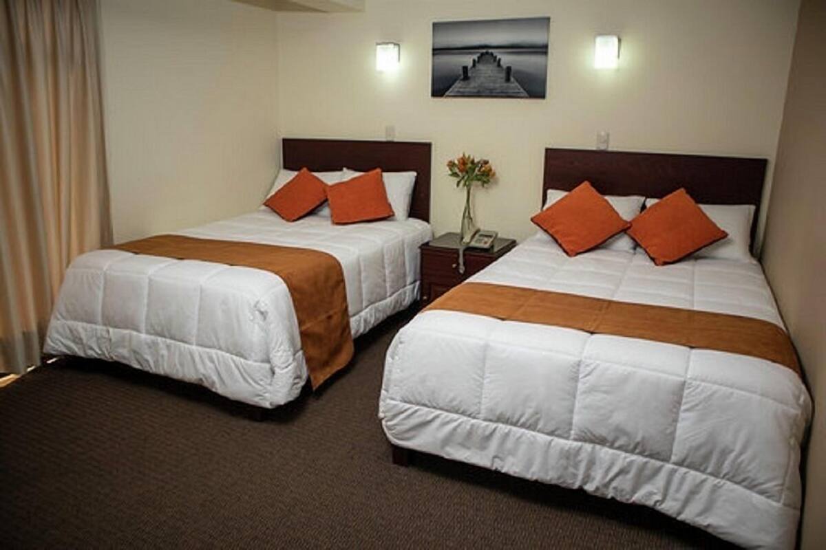 Two double beds with white bedding and orange pillows, a nightstand with a vase of flowers, and a framed picture on the wall.