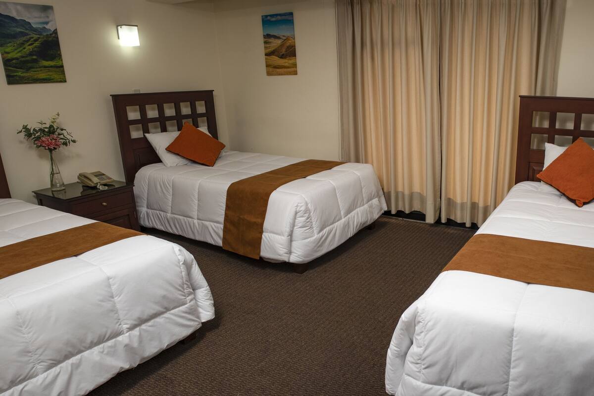 A hotel room with three single beds, each with a wooden headboard, white bedding, and orange pillows. There is a small table with a vase of flowers and a telephone.