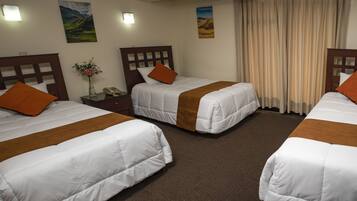 Habitación de hotel con tres camas individuales, cada una con cabecera de madera, ropa blanca y almohadas naranjas. Hay una mesita con un jarrón de flores y un teléfono.