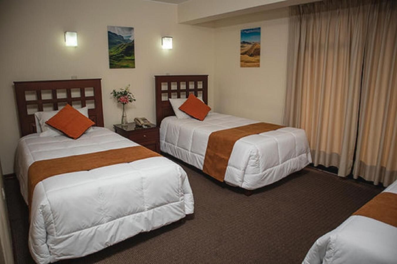 Habitación de hotel con dos camas, cada una con cabecero de madera y ropa de cama blanca. Hay dos luces fijadas a la pared, una mesita de noche con un florero y una ventana con cortinas.