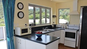 A modern kitchen with white cabinets, a black countertop, and a bowl of fruit. There are two clocks on the wall, a window with a view of greenery, and a stove. - Jewel in Tata Beach. (Tata Beach)